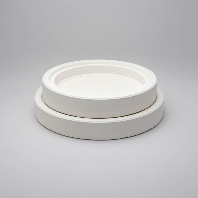 May include: Two stacked circular white ceramic plates with smooth, matte surfaces. The top plate is smaller in diameter than the bottom plate, and both feature a raised, flat-edged rim design that creates a clean, uniform, and solid white appearance.