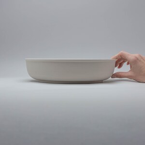 Pode incluir: Uma tigela de cer&acirc;mica cinza claro com formato arredondado. A tigela &eacute; segurada por uma m&atilde;o em um fundo branco liso. A tigela tem aproximadamente 30 cm de di&acirc;metro.