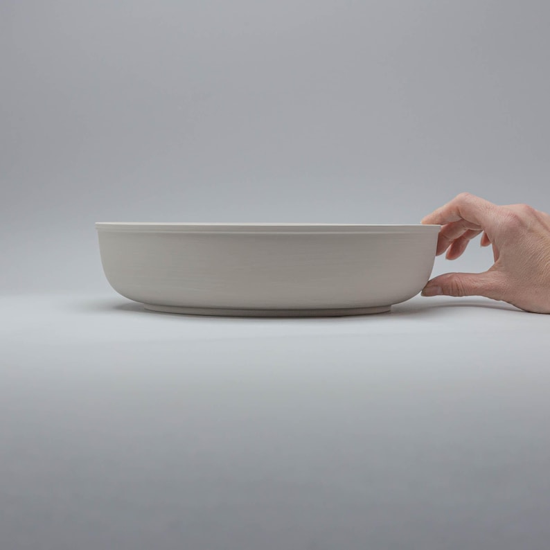 Pode incluir: Uma tigela de cer&acirc;mica cinza claro com formato arredondado. A tigela &eacute; segurada por uma m&atilde;o em um fundo branco liso. A tigela tem aproximadamente 30 cm de di&acirc;metro.