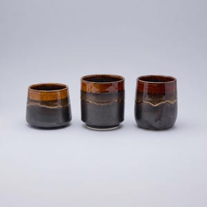 May include: Three ceramic cups with a two-tone glaze. The lower portion of each cup is a matte charcoal black, while the upper rim features a glossy, warm amber and brown drip pattern. The cups vary slightly in height and width, with a smooth finish.