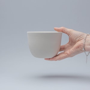 Peut inclure: Un bol en céramique blanc cassé lisse, de forme arrondie. Le bol est tenu par une main sur un fond gris clair. La conception simple du bol suggère qu'il pourrait être utilisé pour servir ou comme pièce décorative.