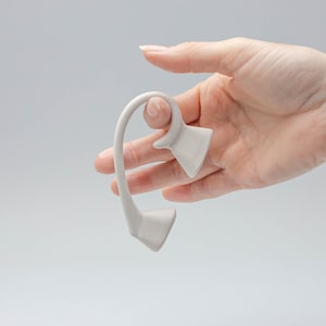 May include: A white ceramic ear cuff with a unique, abstract design. The cuff is shaped like a curved line with a small, rounded protrusion at the top.