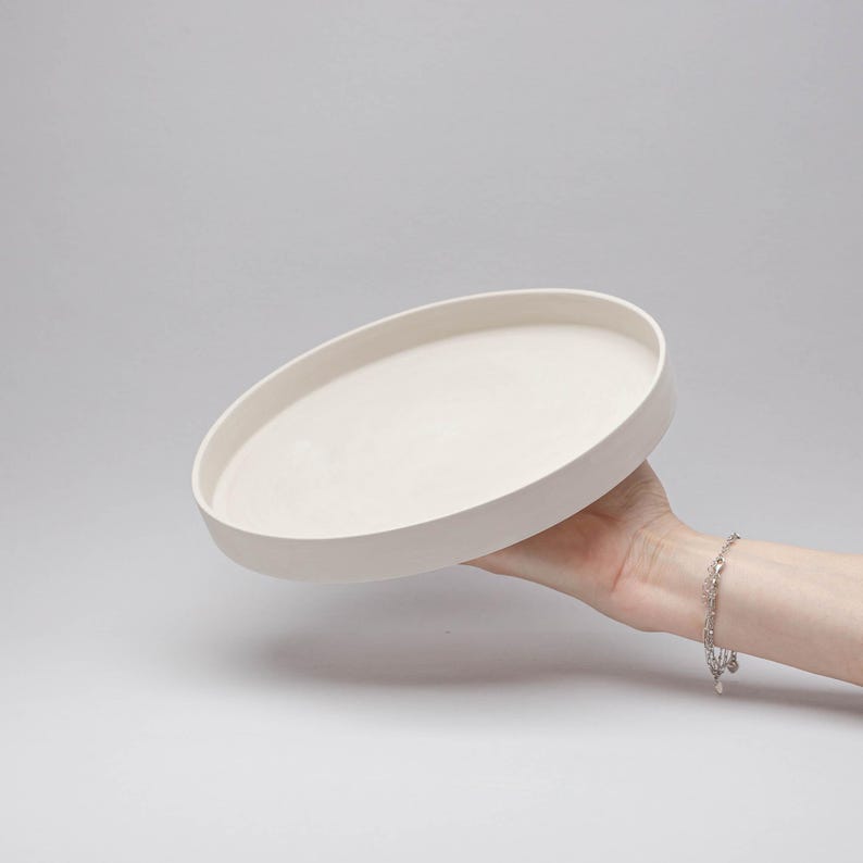 May include: A circular, off-white ceramic tray with a smooth, matte finish and a raised edge. The tray features a flat, level base and a consistent, neutral colour throughout, presenting a simple, functional design for holding various small items.