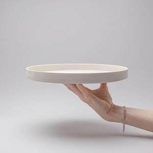 May include: A circular, off-white ceramic tray with a smooth, matte finish and a raised edge. The tray features a flat, level surface and a consistent, neutral colour throughout, presenting a clean and simple design for serving or display purposes.