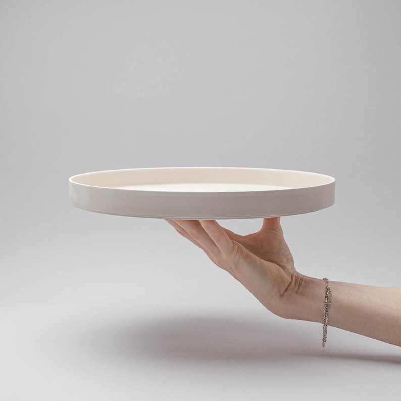 May include: A circular, off-white ceramic tray with a smooth, matte finish and a raised edge. The tray features a flat, level surface and a consistent, neutral colour throughout, presenting a clean and simple design for serving or display purposes.