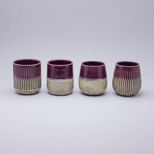 Peut inclure: Quatre gobelets en céramique disposés en rangée. Chaque récipient présente un vernis violet profond sur la partie supérieure et une base crème tachetée. Deux gobelets ont des textures nervurées verticales, tandis que les deux autres sont lisses.