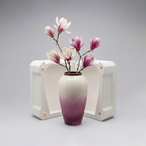 Può includere: Un vaso sfumato bianco e viola che contiene fiori di magnolia con petali bianchi e viola. Il vaso è posto di fronte a una struttura geometrica bianca con schienale aperto e dettagli dorati. I fiori sono in piena fioritura.