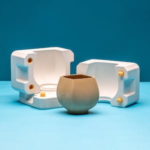 Peut inclure: Un moule en céramique blanche pour faire une tasse en céramique brune. Le moule est en deux parties et a un intérieur lisse et arrondi. La tasse est posée dans le moule, et elle a une base carrée et un sommet arrondi.