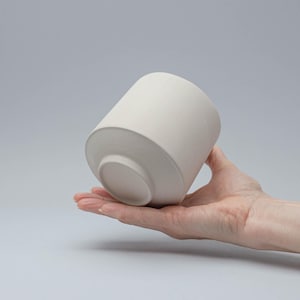 Könnte beinhalten: Ein weißer Keramiktopf mit abgerundetem Boden und leicht ausgestelltem Rand. Der Topf befindet sich in der Hand einer Person.