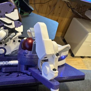 May include: A close-up of a 3D-printed robotic arm component. The arm features a combination of white, purple, and silver parts, with a red sphere at the joint. The base is a textured purple, and the arm has a mechanical design.