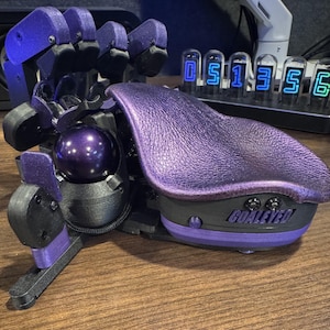 Puede incluir: Una mano robótica en morado y negro, sosteniendo una esfera morada oscura. La mano tiene dedos articulados y un reposamanos morado texturizado. La base de la mano tiene el texto "GOALEYED".