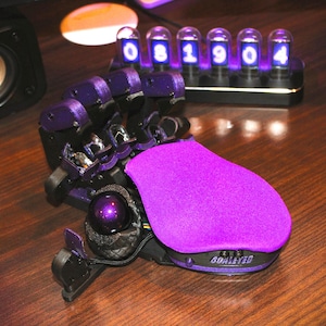 Könnte beinhalten: Eine violette, im 3D-Druck hergestellte Roboterhand mit einer violetten Stoff-Handauflage. Die Hand hat eine schwarze Kugel am Daumenende und eine schwarze Basis mit einem digitalen Display, das "081904" anzeigt.