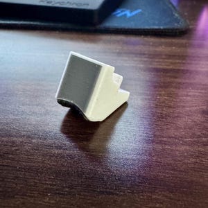 May include: A small, white and black 3D-printed object with a rectangular shape. The object has a stepped design and appears to be a support or stand. The background includes a keyboard and a mousepad on a wooden surface.