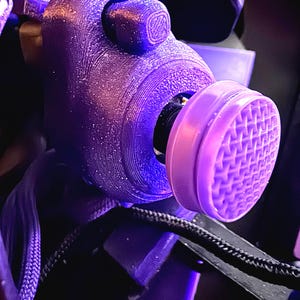 Puede incluir: Una máscara respiratoria morada impresa en 3D con un filtro morado claro y una correa negra. La máscara tiene una superficie texturizada y un cuerpo cilíndrico. El filtro tiene un patrón de rejilla.