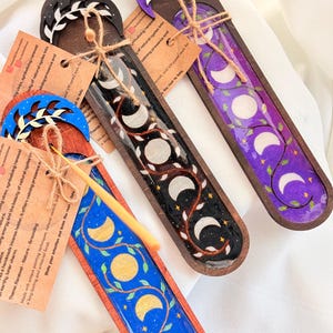 May include: Handmade incense holders in blue, black, and purple, featuring moon phase designs and leaf accents. Each holder is made of wood and has a unique painted design. The holders are tied with twine and have small tags attached.