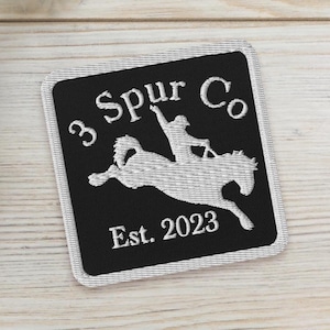 May include: A square, black patch with white embroidered text and a graphic of a person riding a bucking horse. The text reads "3 Spur Co" and "Est. 2023". The patch has a white border and is set against a light wood grain background.