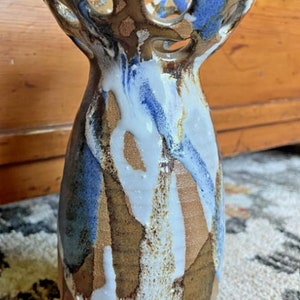 Op de afbeelding: Een keramische vaas met een uniek, abstract ontwerp met bruine, blauwe en witte glazuur. De vaas heeft een brede, gegolfde rand met uitgesneden cirkels.