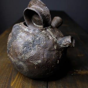 Puede incluir: Una tetera de cerámica marrón oscuro con una superficie texturizada y una tapa con un asa de anillo. La tetera está sobre una superficie de madera.
