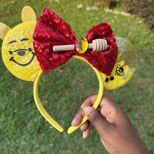 May include: A yellow headband with attached ears and a red sequined bow. The ears are yellow with a smiling face and honey-themed accents. A wooden honey dipper is attached to the bow. The headband is held by a person.