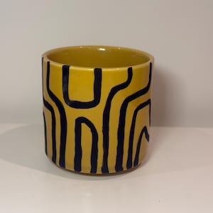 May include: A cylindrical ceramic mug featuring a vibrant mustard yellow base colour. The exterior is decorated with a series of bold, hand-painted black vertical lines that curve and connect in a continuous, abstract pattern around the entire vessel.