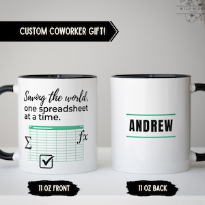 May include: A white ceramic mug with a black handle and rim. The front features the text 'Saving the world, one spreadsheet at a time' with a green spreadsheet grid, a sigma symbol, a checkmark, and an fx symbol. The back displays the name 'ANDREW'.