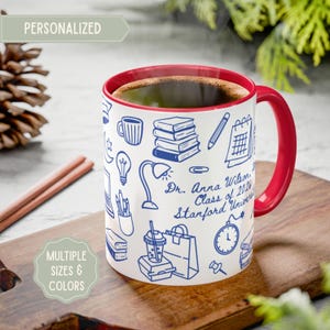 Puede incluir: Taza de cerámica blanca con interior y asa rojos, llena de café. La taza presenta dibujos lineales azules de útiles escolares y el texto "Dr. Anna Wilson, Class of 2026, Stanford University". La taza está etiquetada como "PERSONALIZADA" y "MÚLTIPLES TAMAÑOS Y COLORES".