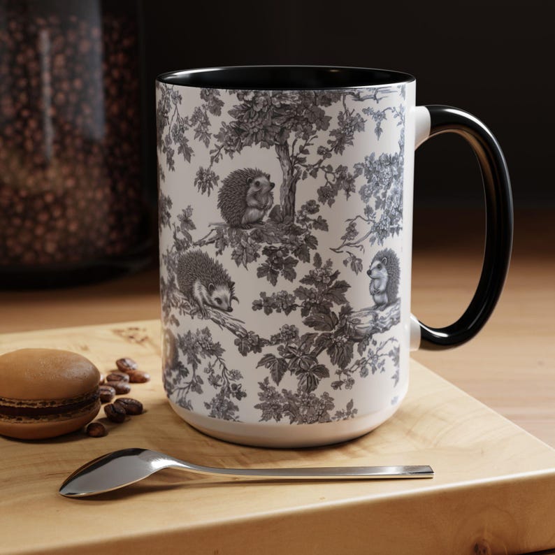 May include: A white ceramic mug with a black interior and handle, featuring a detailed black and white toile-style pattern of hedgehogs perched on tree branches amidst leaves and flowers, standing upright with a smooth cylindrical body.