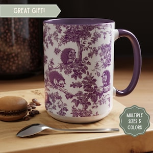 May include: White ceramic mug with a purple interior and handle. The mug features a purple toile design with hedgehogs and foliage. Text on the mug reads "GREAT GIFT!" and "MULTIPLE SIZES & COLORS". A spoon and a macaron are on a wooden surface.