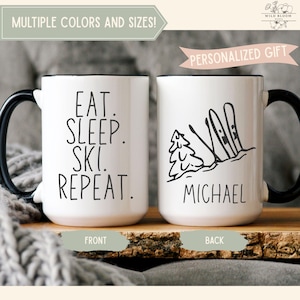 Peut inclure: Deux mugs en céramique blancs avec des poignées et des bords noirs. L'un des mugs porte l'inscription "EAT. SLEEP. SKI. REPEAT." en noir. L'autre mug présente un dessin noir de skis et d'un arbre, avec le nom "MICHAEL" en dessous.