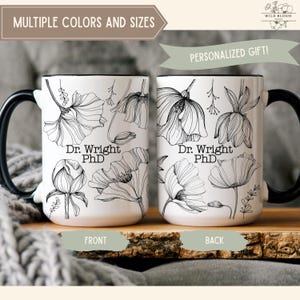 Puede incluir: Dos tazas de cerámica blancas con asas y bordes negros, con arte lineal floral negro y el texto "Dr. Wright PhD." Las tazas están etiquetadas como "Delante" y "Detrás" y se muestran sobre una superficie de madera.