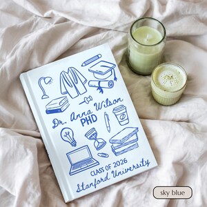 May include: Sky blue graduation journal with blue line art illustrations of academic symbols. The journal is personalized with the name "Dr. Anna Wilson, PHD", "Class of 2026", and "Stanford University". A green candle and drink are next to the journal.