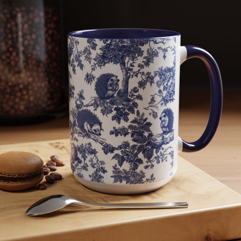 May include: A white ceramic mug featuring a blue toile-style pattern of hedgehogs perched on tree branches. The mug has a solid dark blue interior and a matching dark blue handle. The exterior surface is decorated with detailed illustrations of hedgehogs.