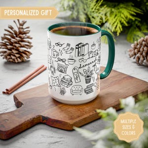 Peut inclure: Une tasse en céramique blanche avec une anse et un bord verts, remplie d'un liquide foncé. La tasse est décorée de dessins au trait noirs de livres, de lunettes et d'autres objets confortables. Le texte sur la tasse indique "Savannah". Les mots "CADEAU PERSONNALISÉ" et "PLUSIEURS TAILLES ET COULEURS" sont également visibles.