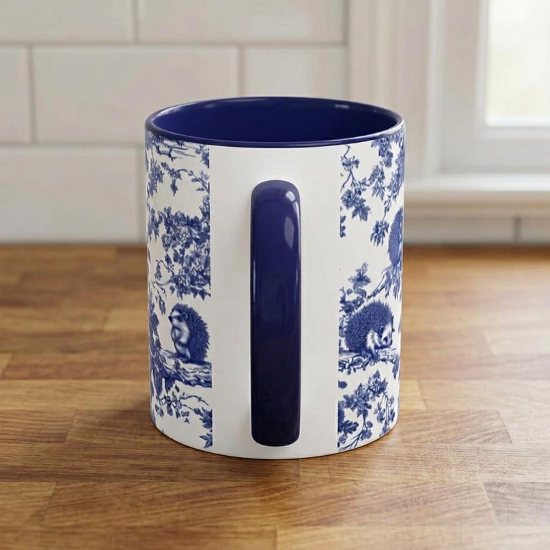 May include: A white ceramic mug featuring a dark blue handle and interior. The exterior surface is decorated with a repeating blue and white toile pattern depicting hedgehogs perched on tree branches amidst detailed floral and leafy foliage.