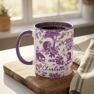 May include: A white ceramic mug with a purple handle and interior, featuring a purple toile-style pattern of hedgehogs among trees and foliage. The name Charlotte is printed in a cursive script on the front of the mug, which is filled with coffee.