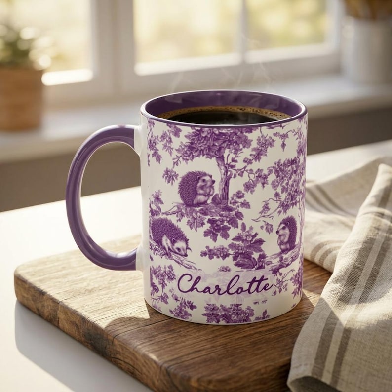 May include: A white ceramic mug with a purple handle and interior, featuring a purple toile-style pattern of hedgehogs among trees and foliage. The name Charlotte is printed in a cursive script on the front of the mug, which is filled with coffee.