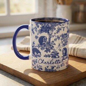 May include: A white ceramic mug with a dark blue handle and a blue toile-style pattern featuring hedgehogs among trees and foliage. The name Charlotte is printed in a blue cursive script on the front of the mug, which is filled with dark coffee.