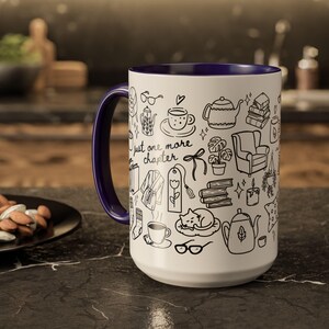 May include: White ceramic mug with a dark blue interior and handle. The mug is decorated with black line drawings of books, teapots, and a cat. The text "Just one more chapter" is also featured on the mug.