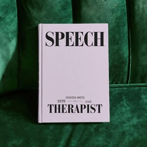 May include: A light lavender hardcover book with black text on the front cover. The text reads SPEECH at the top and THERAPIST at the bottom, with JESSICA SMITH and ESTD 2026 printed in the center. The book has a clean, minimalist design.