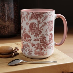 May include: A ceramic mug with a light pink interior and handle, featuring a white exterior decorated with a repeating red toile pattern of hedgehogs perched on tree branches among leaves and flowers, creating a detailed woodland scene.