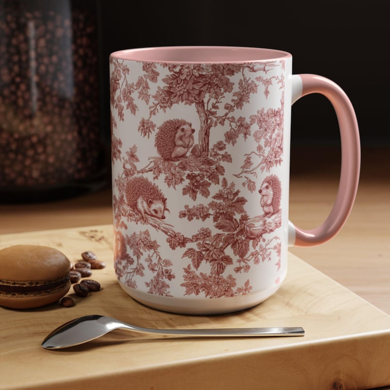 May include: A ceramic mug with a light pink interior and handle, featuring a white exterior decorated with a repeating red toile pattern of hedgehogs perched on tree branches among leaves and flowers, creating a detailed woodland scene.