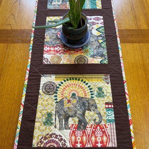 May include: A quilted table runner with a brown border and colorful patterned fabric. The center features three rectangular sections with a repeating elephant design and other decorative patterns. A small potted plant sits on top.