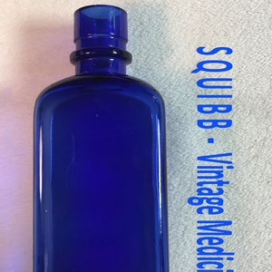 May include: A cobalt blue glass medicine bottle with a black plastic stopper. The bottle is rectangular with rounded edges. The text "SQUIBB - Vintage Medicine Bottle" is written in blue on a white background.