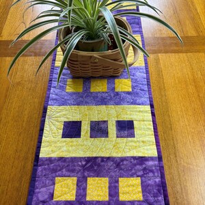 May include: A handmade quilted table runner in shades of purple and yellow. The runner features a repeating pattern of squares and a floral design. A woven basket with a green plant sits on top of the runner.