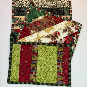 May include: A stack of Christmas-themed quilted placemats. The placemats feature various patterns, including holly, poinsettias, and swirling designs, in shades of red, green, and cream. Each placemat has a green border.