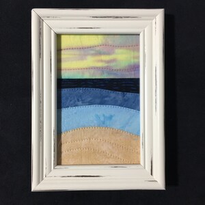 May include: A framed fabric art piece depicting a sunset over a beach scene. The sky is a gradient of yellow, pink, and purple. The ocean is a deep blue with a lighter blue wave. The beach is a light brown.