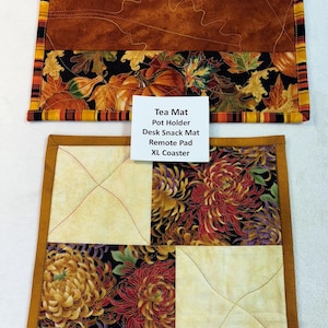 May include: Two quilted mats with autumn-themed patterns. The top mat features a pumpkin and leaf design with a brown center. The bottom mat has a floral pattern with a tan border. Text on a white card reads: Tea Mat, Pot Holder, Desk Snack Mat, Remote Pad, XL Coaster.