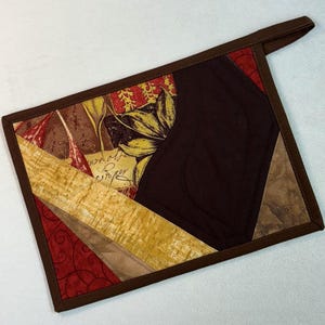 May include: A quilted potholder with a brown border and hanging loop. The design features a patchwork of fabrics in shades of brown, gold, and red, with floral and script patterns. The center is a solid dark brown.