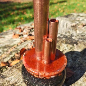 Peut inclure: Une sculpture cuivrée avec quatre tubes cylindriques de hauteurs variables, émergeant d'une base ronde brun rougeâtre. La base repose sur une forme arrondie, sombre et mouchetée. La sculpture est à l'extérieur, sur une surface en bois vieilli.