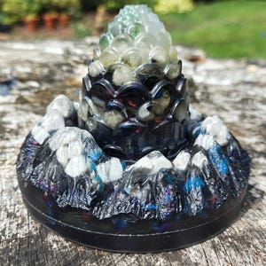 Könnte beinhalten: Dekorative Kunstharzskulptur mit einem Bergketten-Sockel und einer geschichteten zentralen Struktur. Der Sockel ist schwarz, mit weißen und blauen Bergdetails. Die Spitze ist durchscheinend und geschichtet. Das Design ist künstlerisch und detailliert.